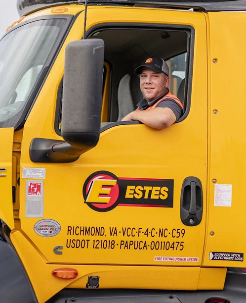 A driver in the cab of a bright yellow Estes freight truck, smiling through the open window.