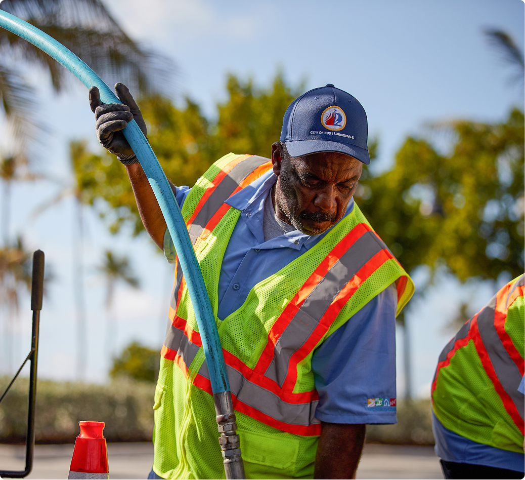 City worker in safety vest and Fort Lauderdale cap handling blue water hose during outdoor maintenance work.