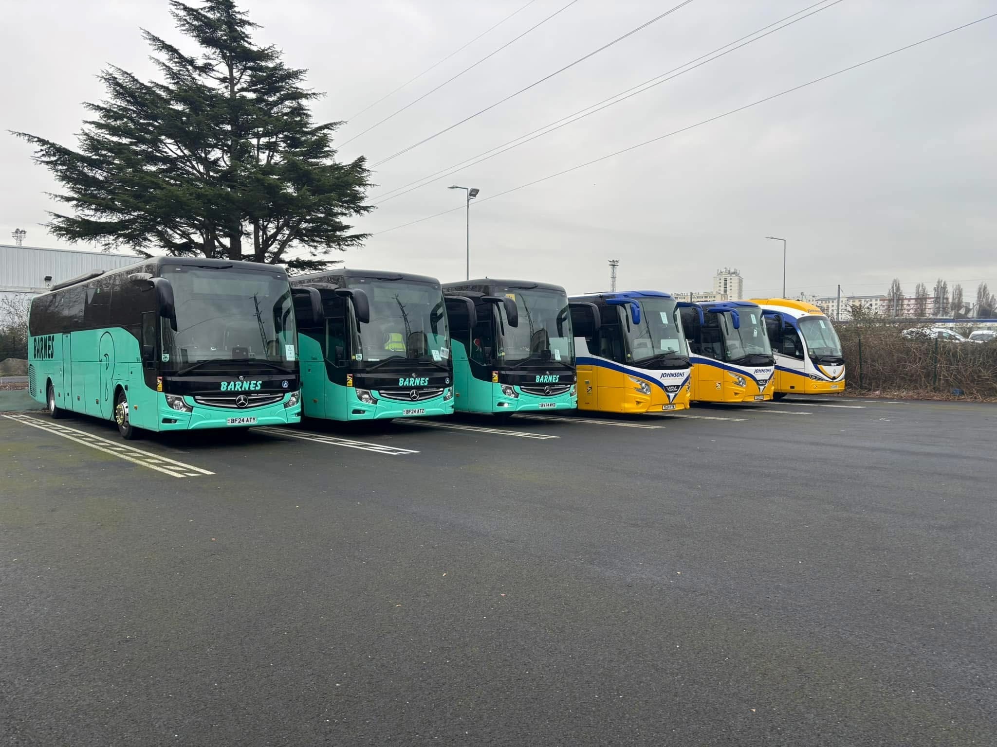 Six coaches parked in a row, three teal Barnes coaches and two yellow and blue Johnsons coaches, in an overcast car park.