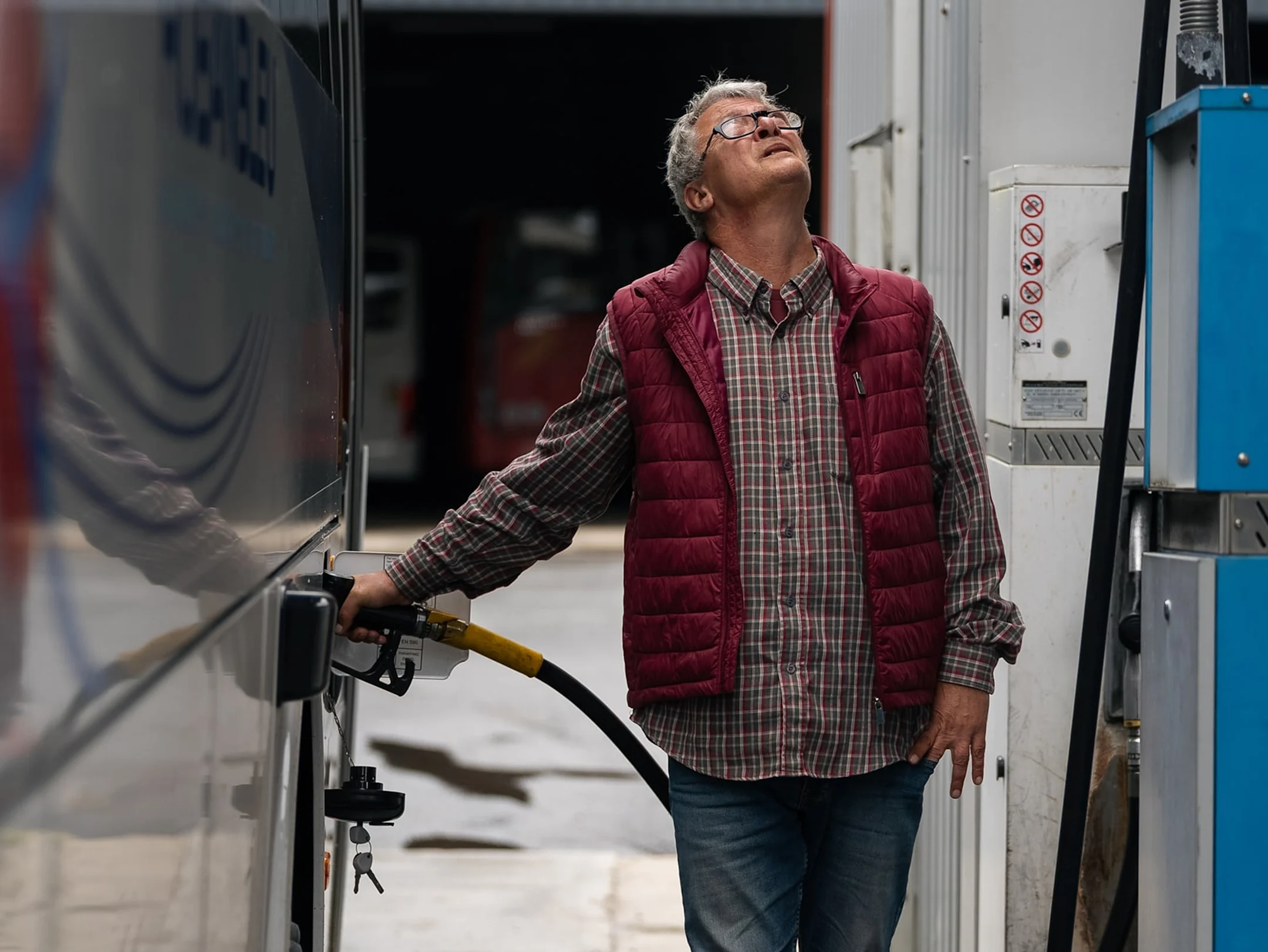 An older man in a red vest and plaid shirt pumps fuel into a large vehicle at a gas station, looking upward.
