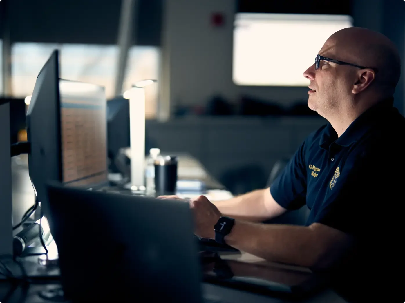Personne avec des lunettes en uniforme de marine sur un ordinateur dans un bureau peu éclairé et une smartwatch au poignet.