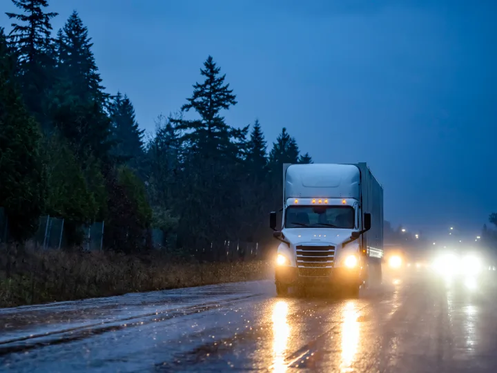 Un camión circula por una carretera mojada bajo un cielo oscuro y nublado, con los faros encendidos y reflejos visibles en el pavimento. Los árboles se alinean al costado de la carretera. 3 Tips de seguridad de flotas en esta temporada de fiestas, Samsara Mexico