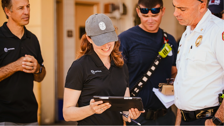 Woman in gray cap smiles while holding a tablet, discussing with a fire official and two others in black uniforms.