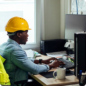Construction professional in yellow hard hat working at computer desk with safety vest and coffee mug nearby.