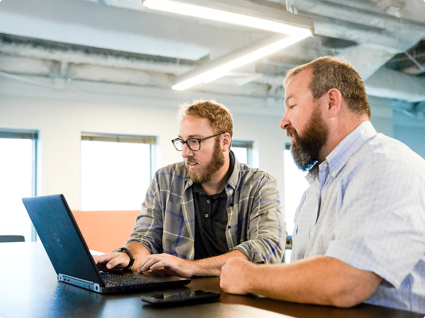 Two bearded men collaborating on a laptop in a bright office space with windows and exposed ceiling.