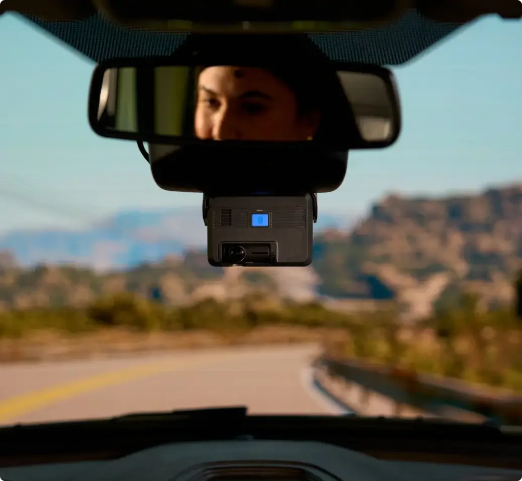View from inside car showing rearview mirror with reflection and dashboard camera mounted below on scenic road.