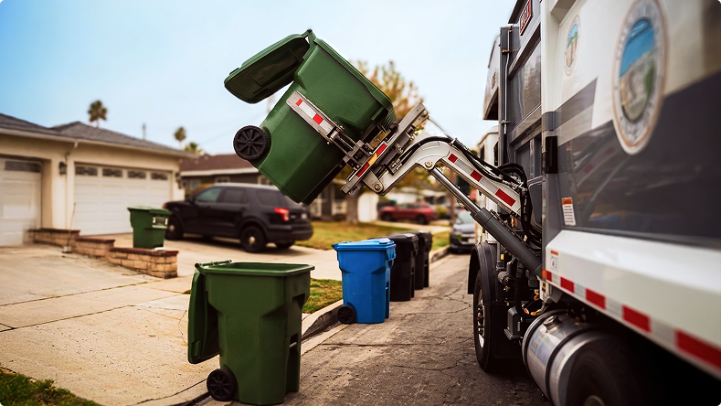 Garbage truck lifting a green bin in a residential neighborhood with blue and black bins on the curb.