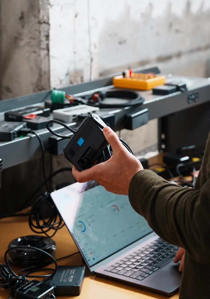 Hand holding electronic device at workbench with laptop and various tools in industrial workshop setting.