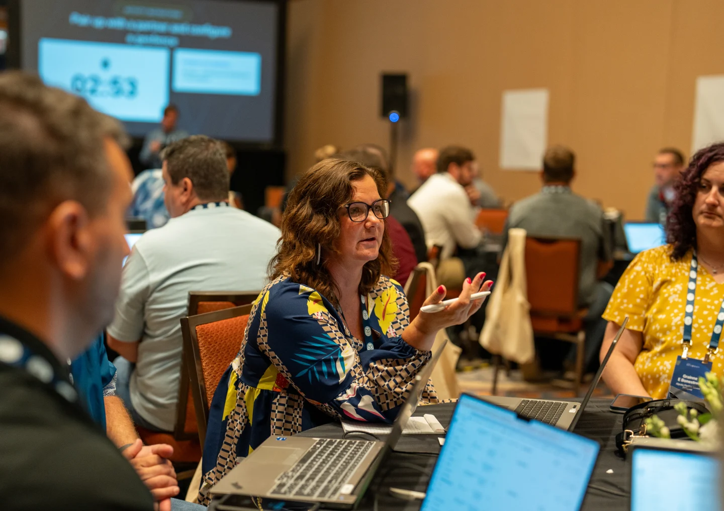 A woman speaks in a group discussion at a conference. Laptops and participants are visible around the table.