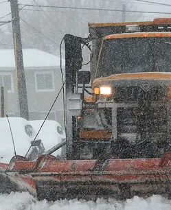 Snow plow truck clearing a road during heavy snowfall with headlights on and operator visible in cab.