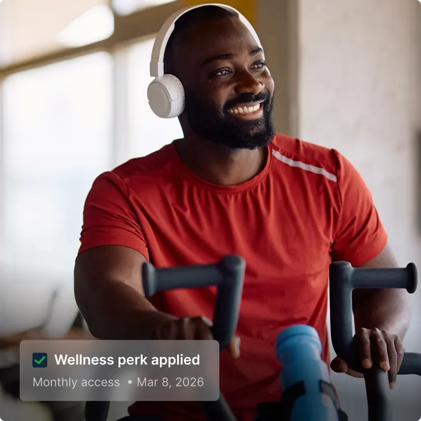 Person in red shirt wearing white headphones exercising on gym equipment with "Wellness perk applied" notification visible.