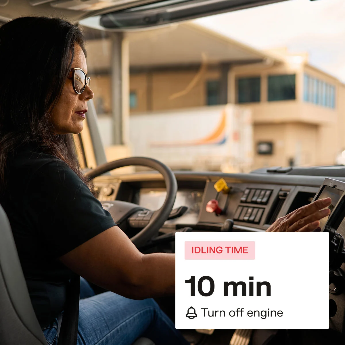 Female truck driver in cab checking dashboard tablet, with an idling time alert showing 10 min and turn off engine warning.