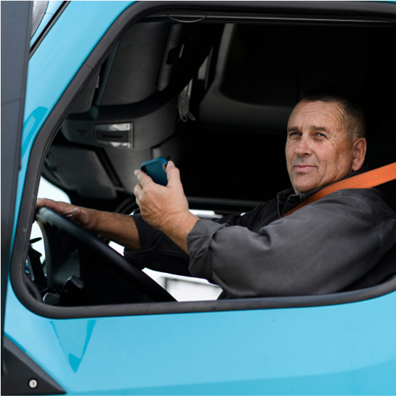 A man in a blue truck's driver seat holds a device while wearing a seatbelt, looking towards the camera.