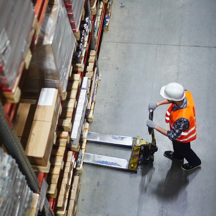 Warehouse worker in orange vest and white hard hat operating a pallet jack near stacked shelves.