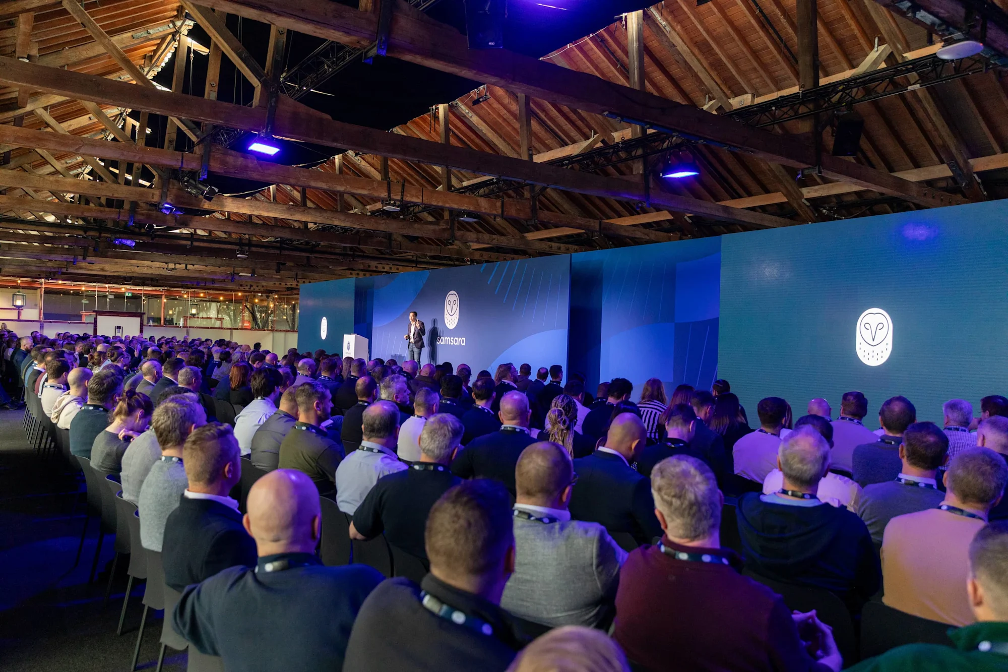 Conferentie in houten zaalruimte met groot publiek luisterend naar spreker op podium met blauw verlichte Samsara-branding.