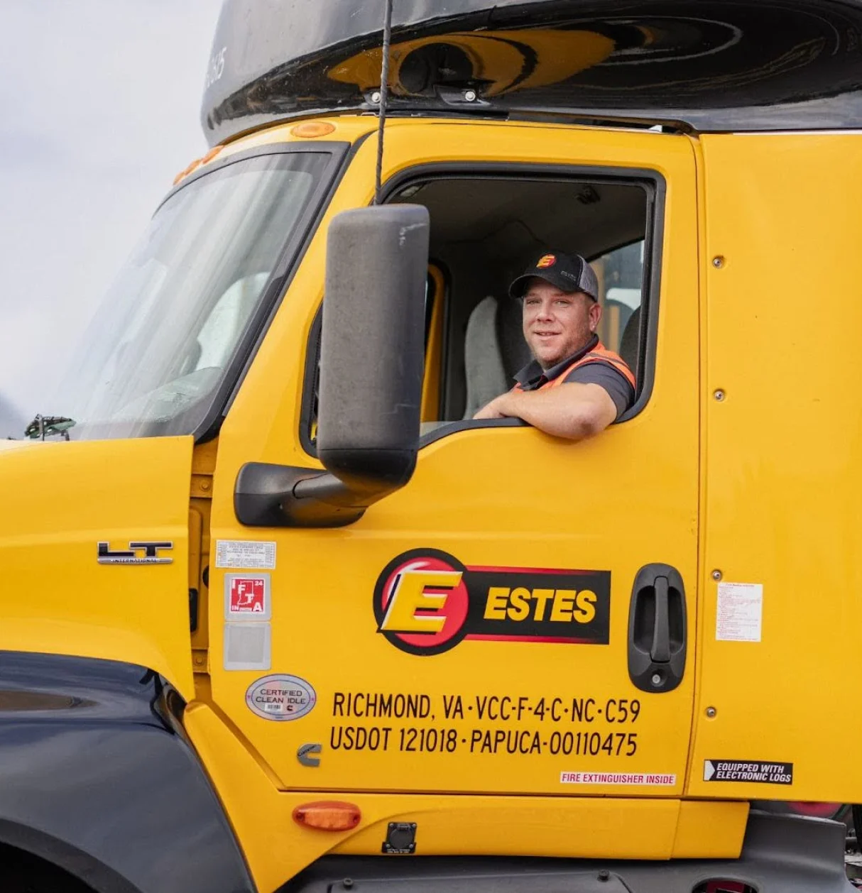 Smiling truck driver in Estes delivery vehicle wearing safety vest, yellow truck cab with Estes logo visible. 