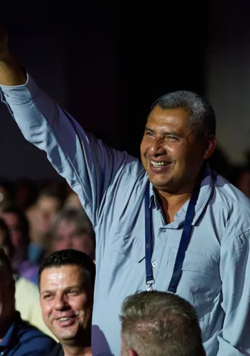 A smiling man in a light blue shirt raising his arm in celebration at a crowded event with audience members visible below.