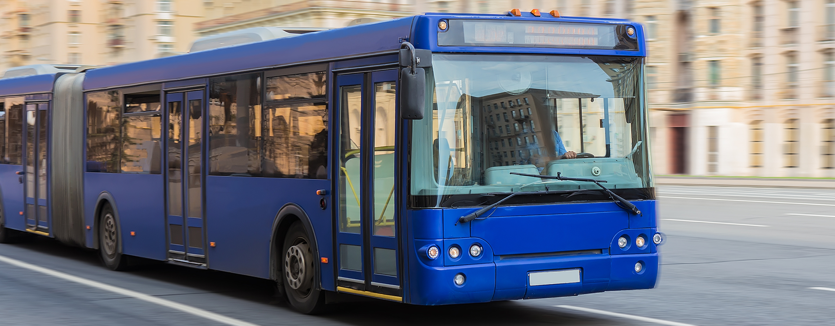 Blue city bus driving through an urban area with apartment buildings in the background.