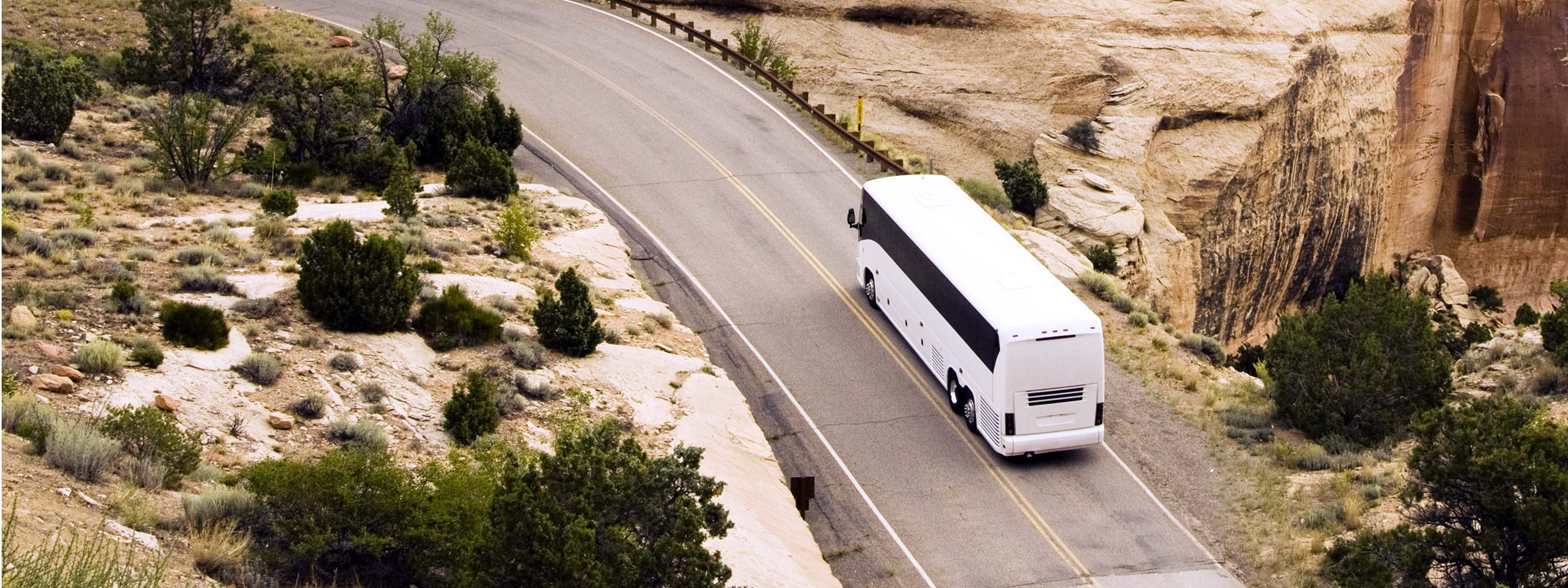 White tour bus driving along a winding desert road with rocky cliffs and sparse vegetation in the American Southwest.