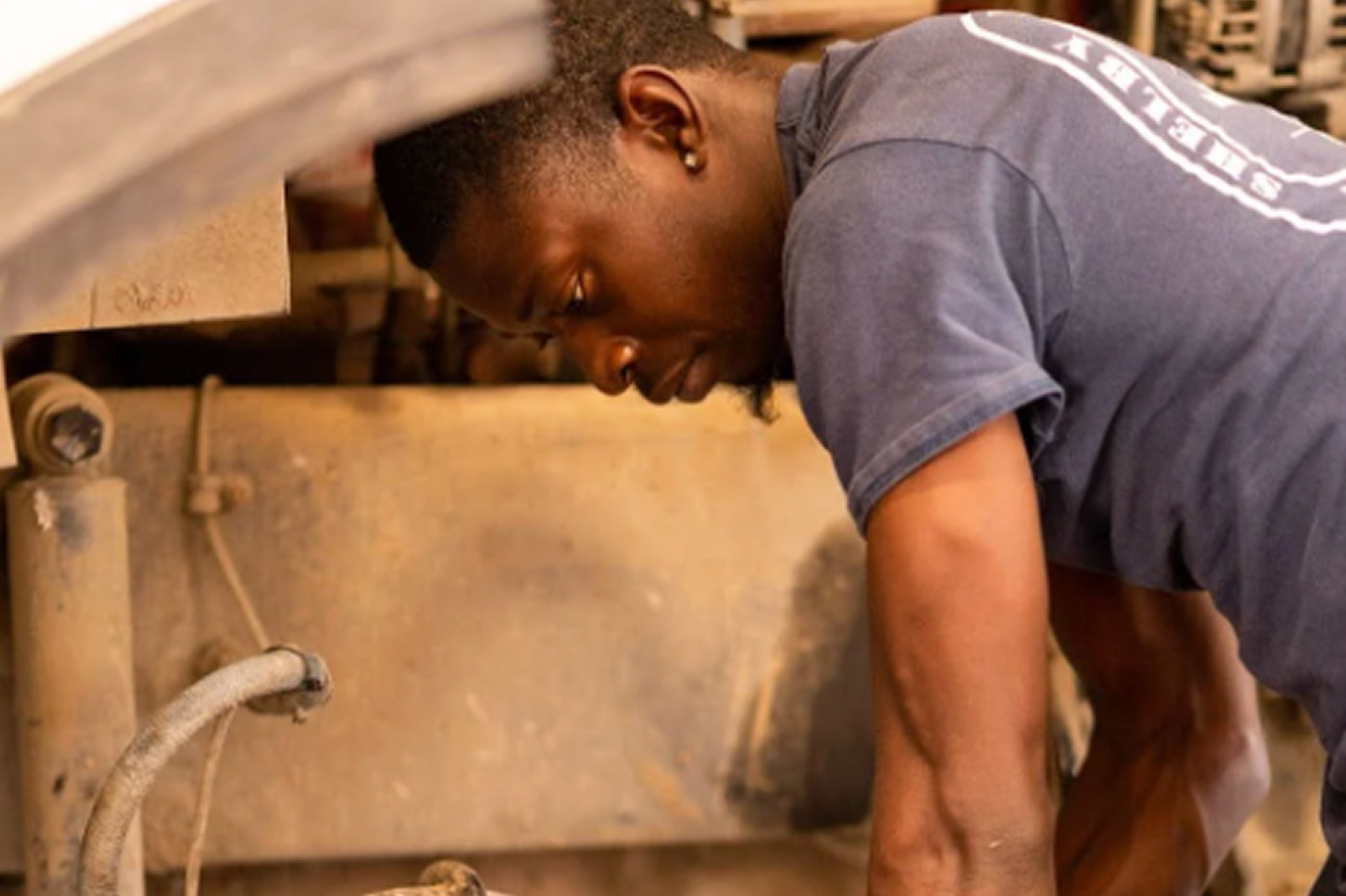 Mechanic in blue shirt working on vehicle undercarriage with visible wheel hub and engine components.