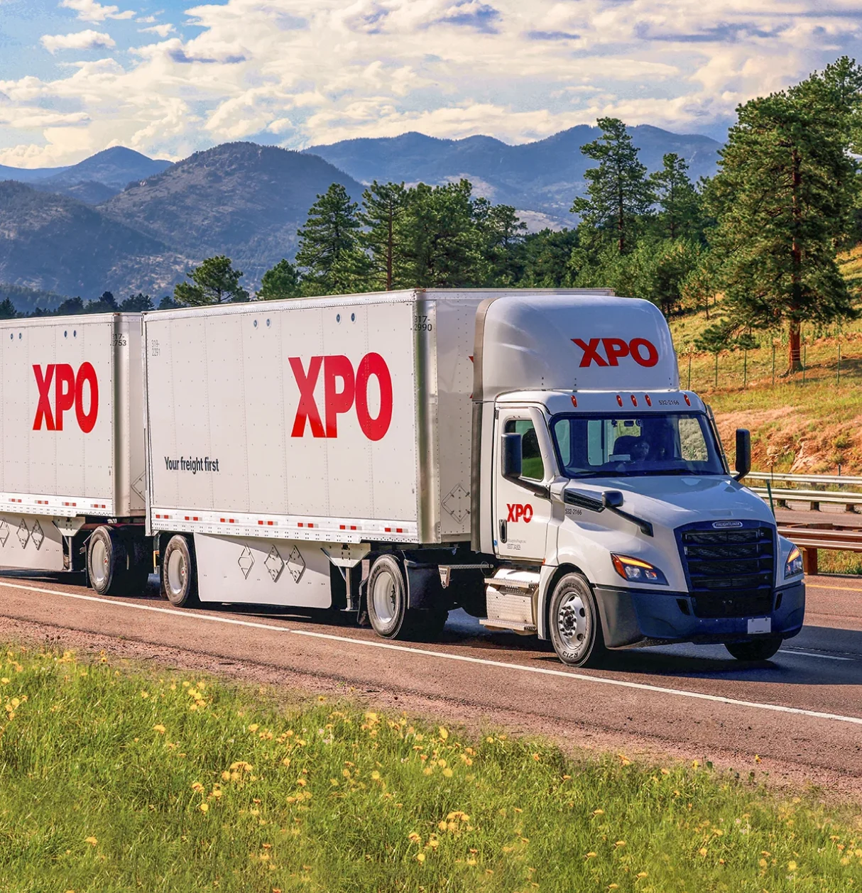 An XPO semi truck drives down a highway with mountains and pine trees in the background.