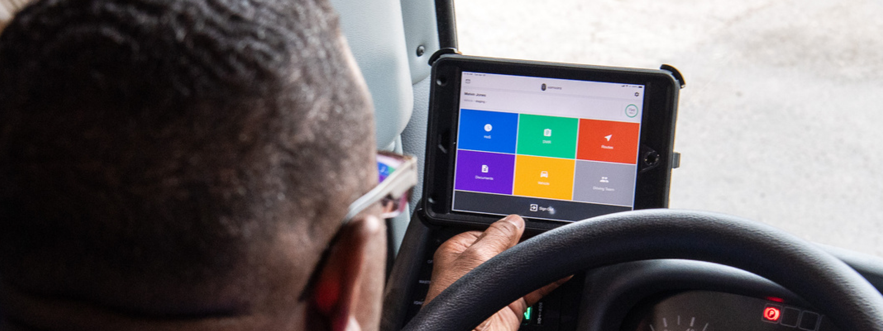 A driver views a mounted tablet displaying a colorful app dashboard with icons for routes, documents, and vehicles.
