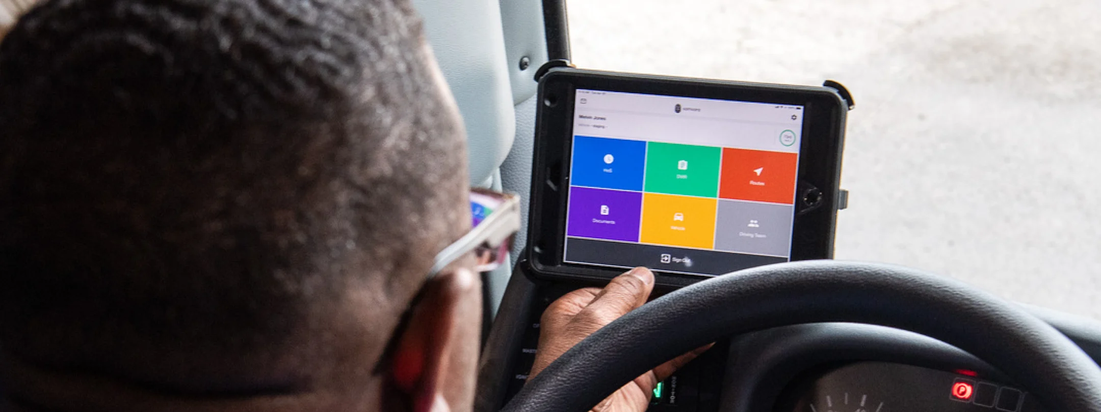 A driver views a mounted tablet displaying a colorful app dashboard with icons for routes, documents, and vehicles.