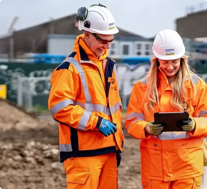 Construction workers in bright orange safety gear and white helmets reviewing information on a tablet at a worksite.