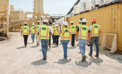 Construction workers in bright safety vests and hard hats walking through an industrial site with equipment and containers.