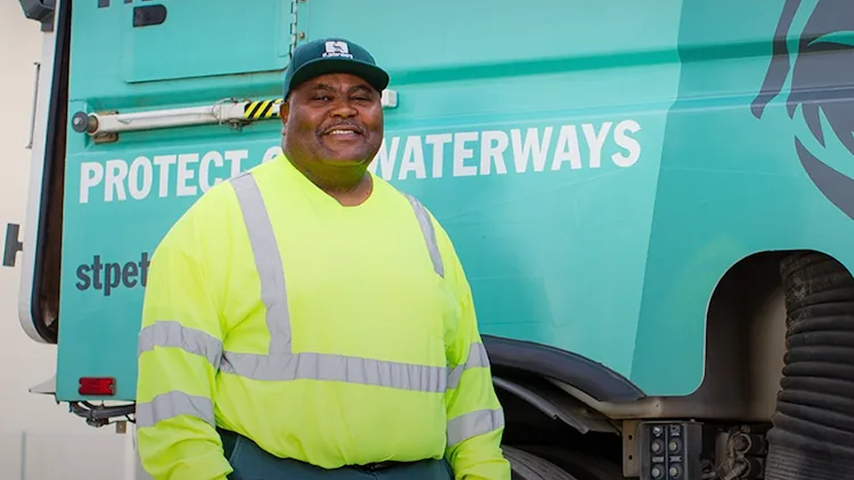 A smiling sanitation worker in a yellow safety vest stands before a teal truck reading "Protect Our Waterways."