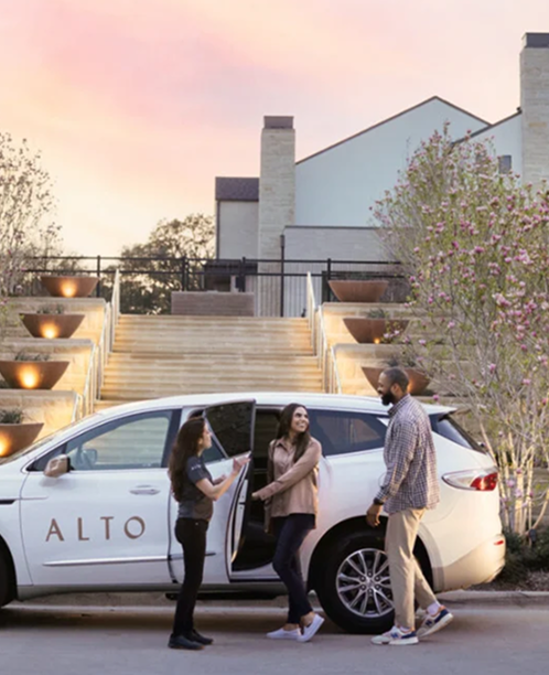 People exiting a white car with "ALTO" logo outside a modern building with illuminated steps at sunset