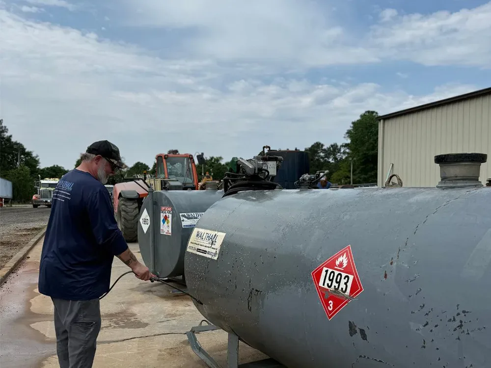 A worker in a blue shirt tends to a large gray fuel tank labeled 1993, with a tractor and warehouse in the background.