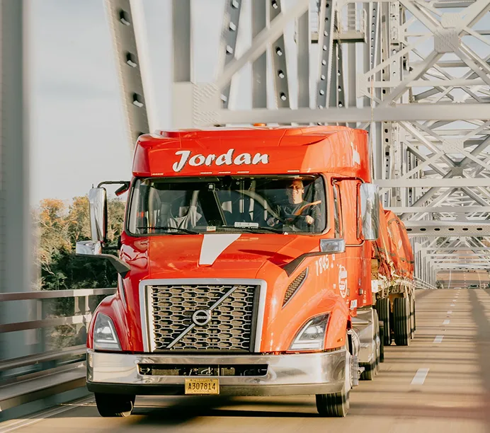 Bright orange Volvo semi-truck with "Jordan" logo crossing a steel bridge structure, driver visible in cab.