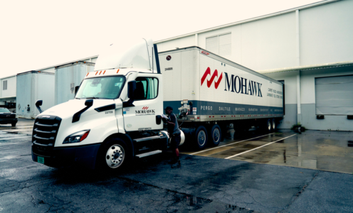 White Mohawk semi-truck with trailer parked at a loading dock on a wet pavement after rain.