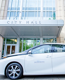 White car parked in front of a modern City Hall building with glass entrance and architectural details.