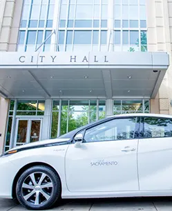 White car parked in front of a modern City Hall building with glass entrance and architectural details.