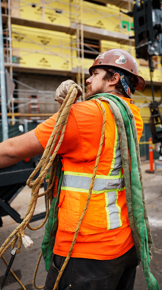 Construction worker in orange safety vest and red hard hat carrying ropes at a building site.