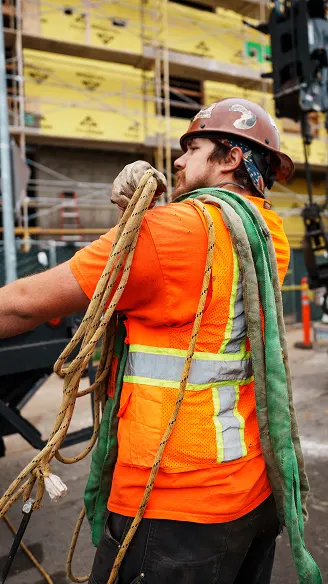 Construction worker in orange safety vest and red hard hat carrying ropes at a building site.