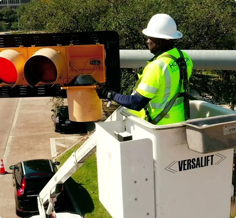 Worker in bright yellow safety vest and white hard hat repairing a traffic light from a bucket lift truck.