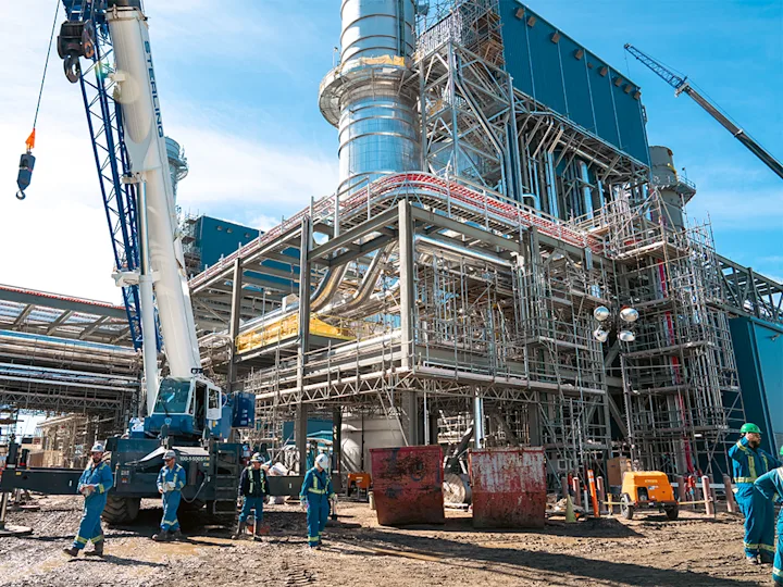 Des ouvriers du bâtiment et une grue dans une installation industrielle avec des échafaudages et des structures métalliques.