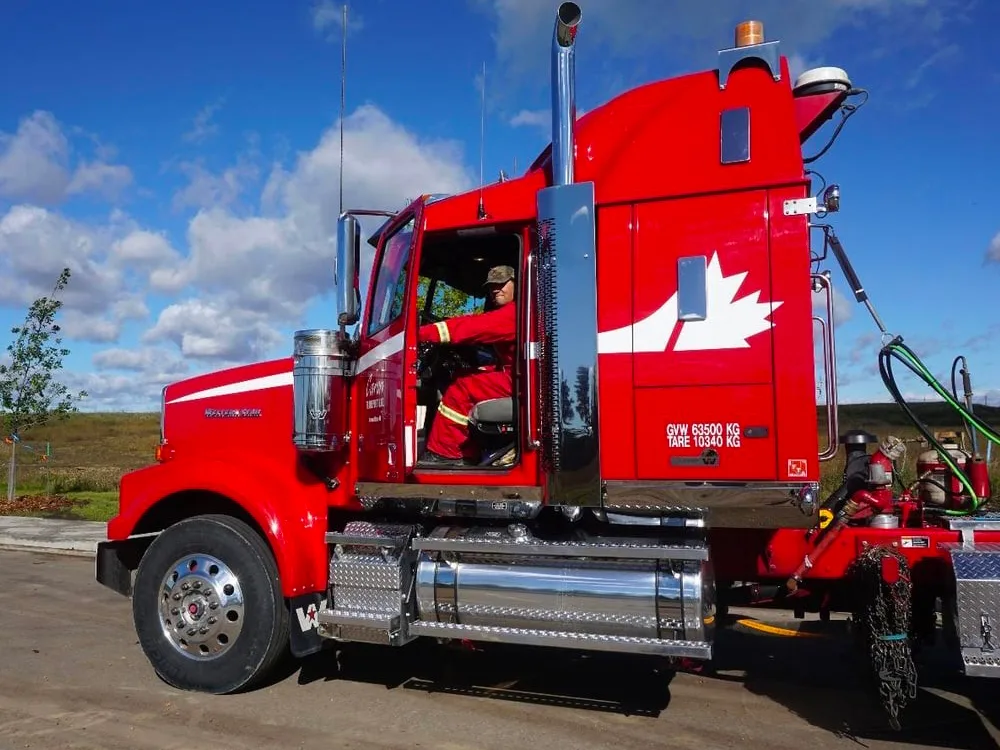 Bright red semi truck with Canadian maple leaf logo parked under blue sky, driver visible in cab.