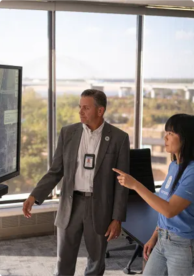 Two professionals in an office with large windows discussing information on a monitor; city view in background.