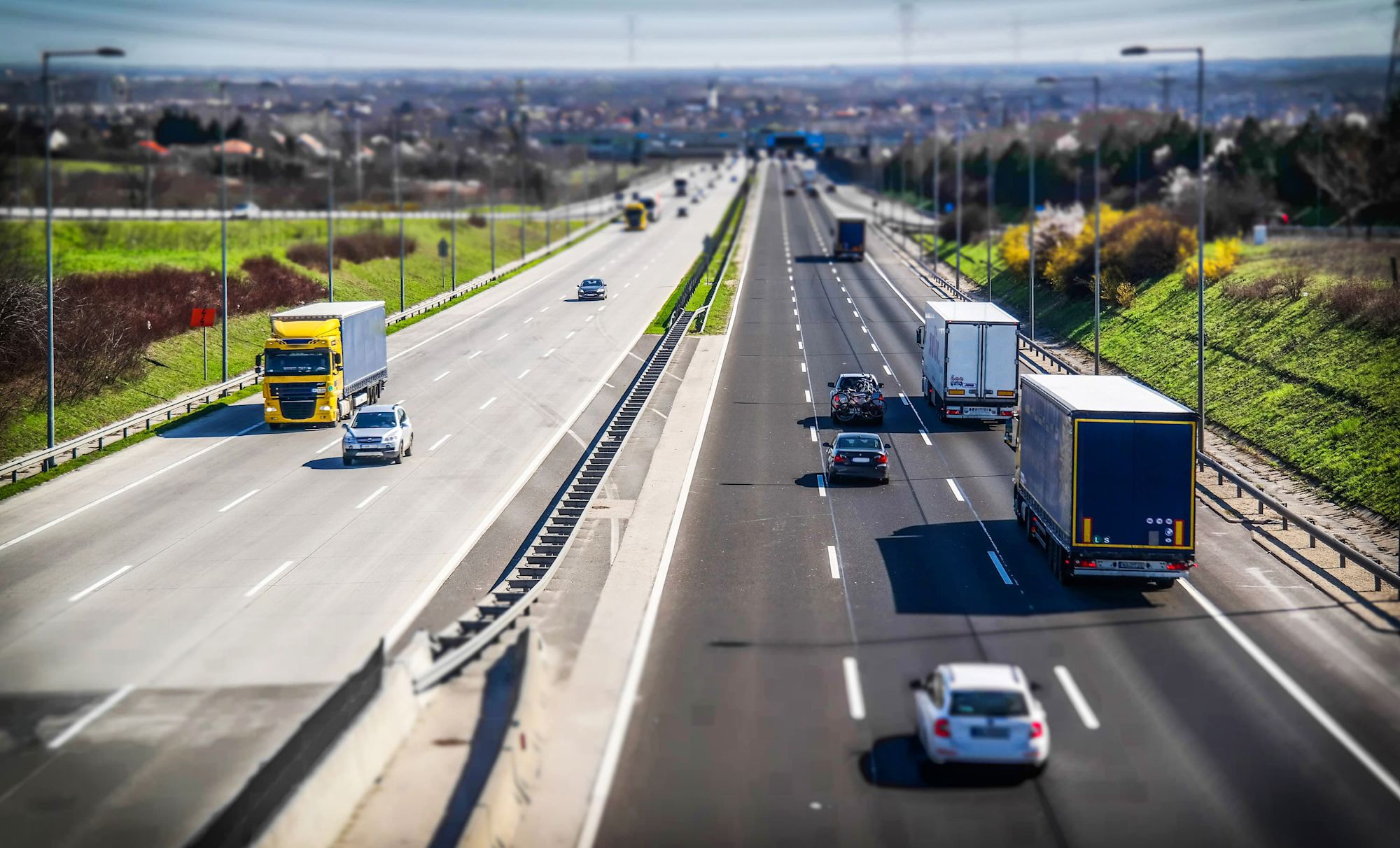 Autobahn mit LKWs und PKWs in beide Richtungen fahrend, umgeben von grünen Feldern und einer Stadt im Hintergrund.