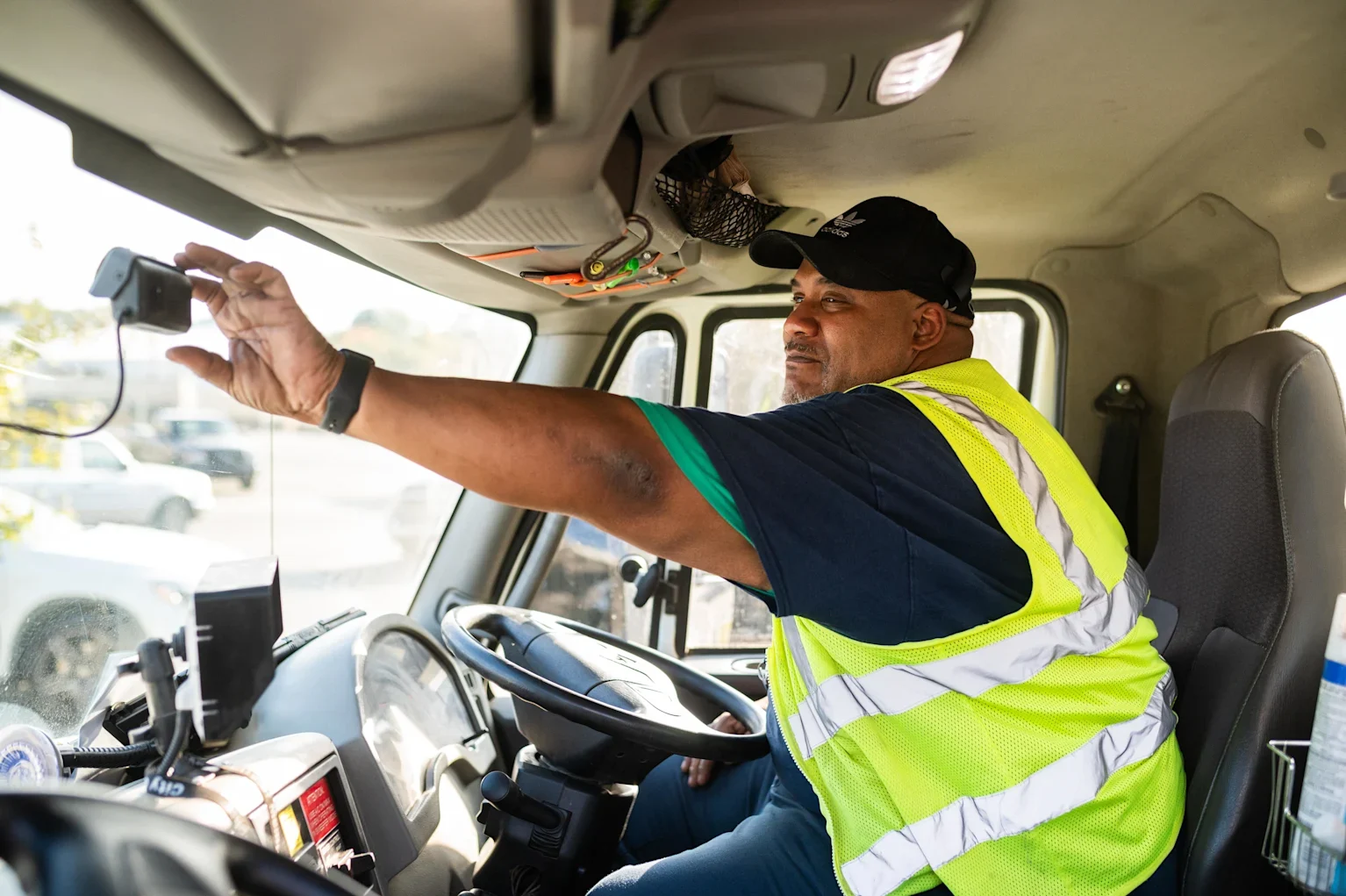 A man in a safety vest adjusts a dash cam while sitting in the driver's seat of a vehicle.