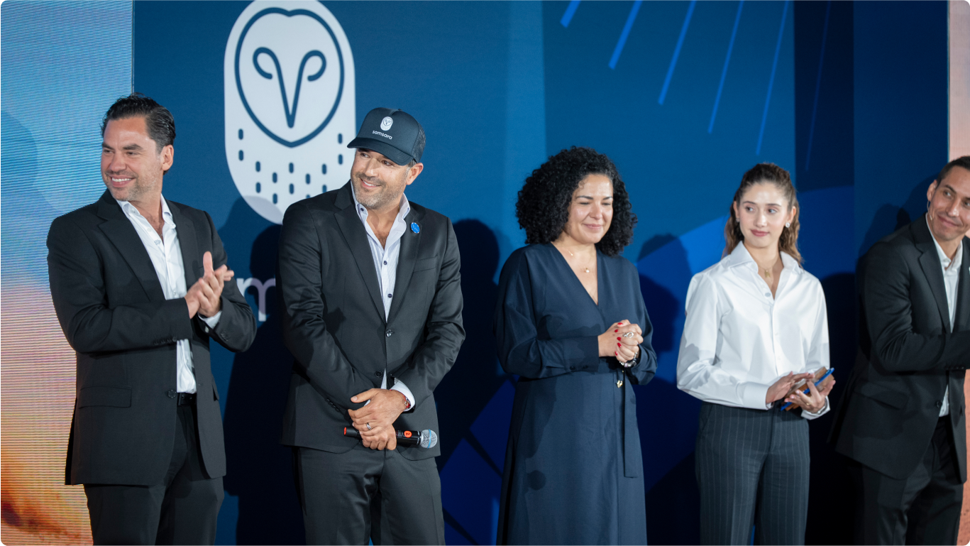 Grupo de personas en traje formal en un evento corporativo con fondo azul y logo de búho blanco de Samsara.