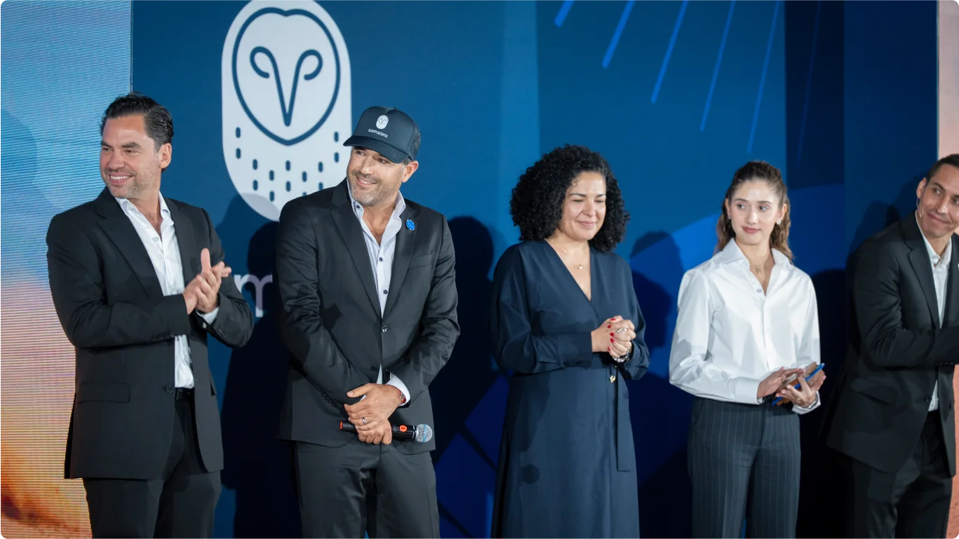 Grupo de personas en traje formal en un evento corporativo con fondo azul y logo de búho blanco de Samsara.