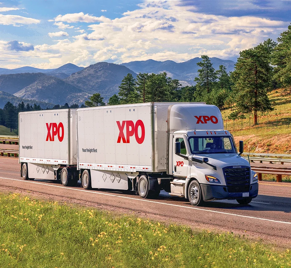 XPO semi-truck with double trailers driving on mountain highway with pine trees and mountain range in background.