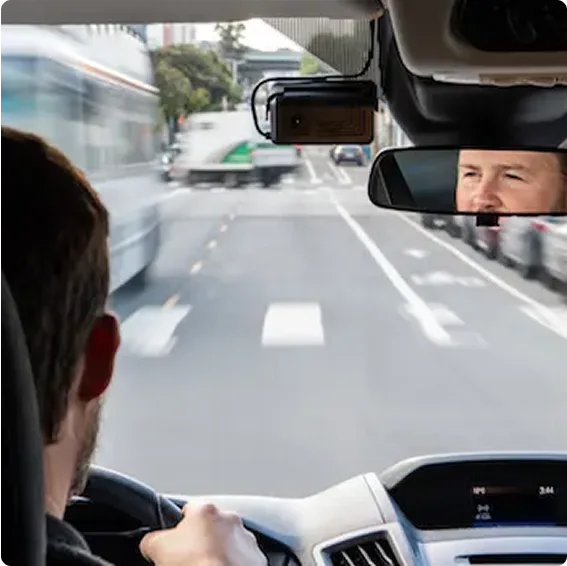 View from inside a car showing the driver's perspective of a city street with a reflection in the rearview mirror.