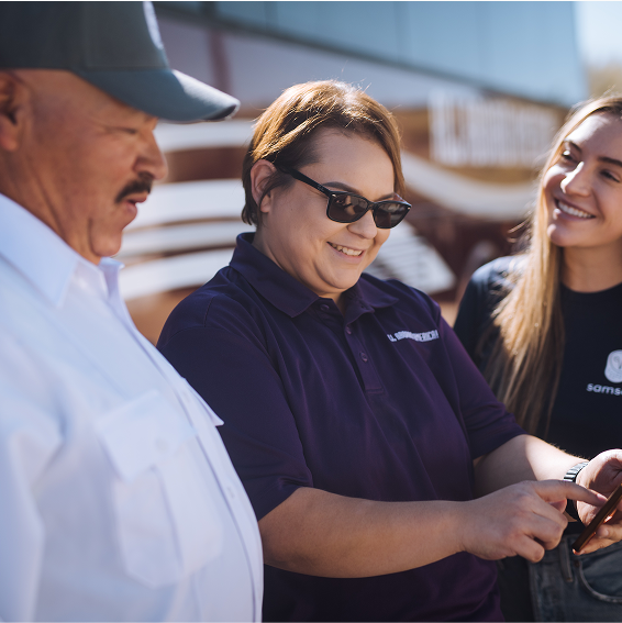 Three people are standing outdoors. A person in sunglasses is holding a device, while the others are engaged in conversation.