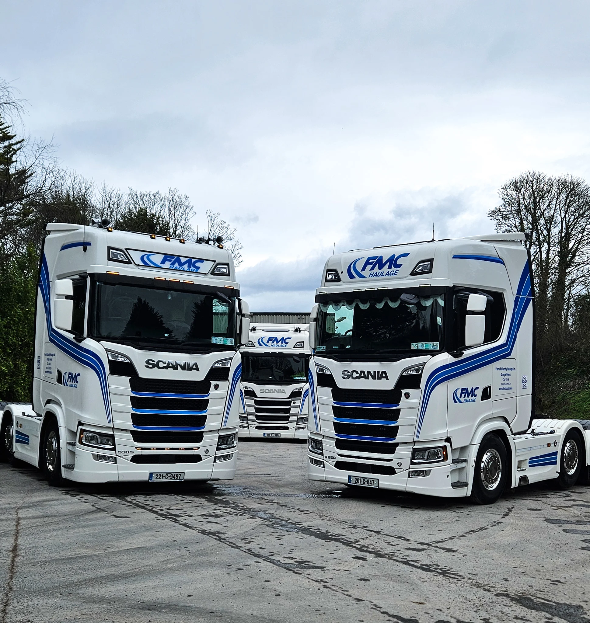 Three white Scania trucks with blue FMC Haulage livery parked in a yard under a cloudy sky.